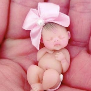 May include: A tiny, sleeping baby doll with a pink bow and a pearl on top of the bow. The doll is made of a soft, realistic material and is posed in a curled-up position.