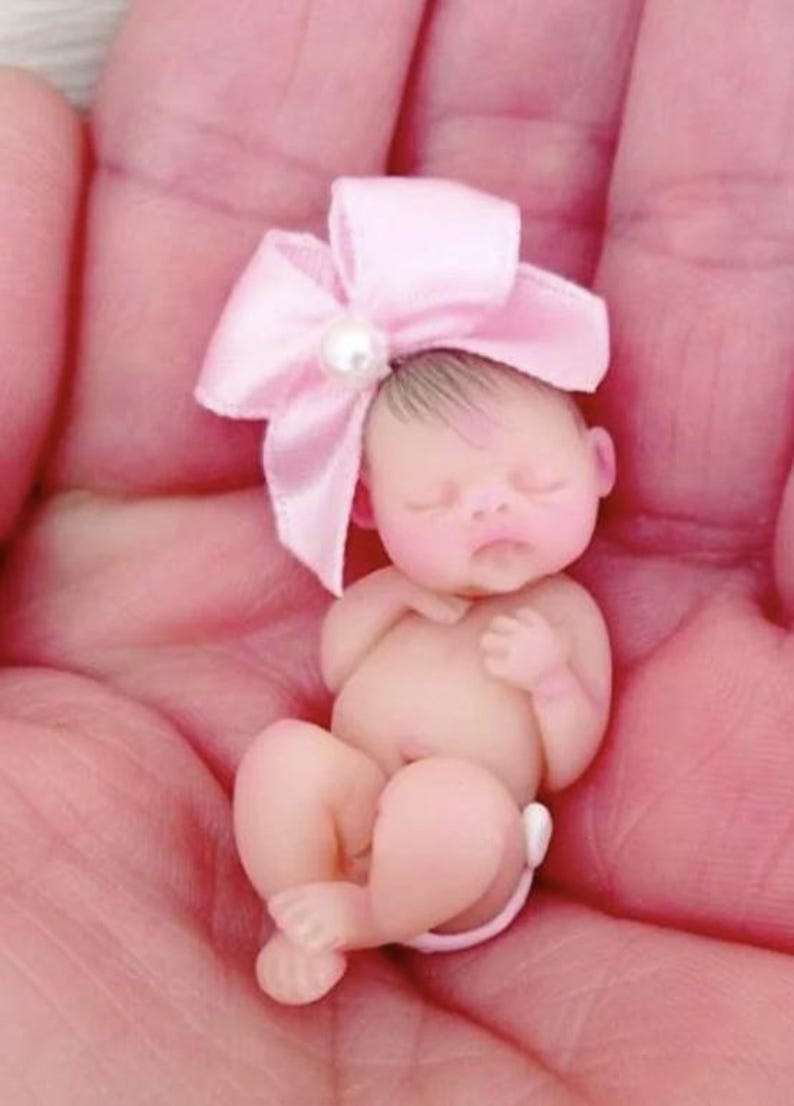 May include: A tiny, sleeping baby doll with a pink bow and a pearl on top of the bow. The doll is made of a soft, realistic material and is posed in a curled-up position.