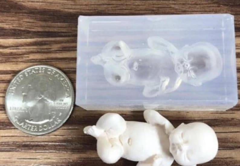 May include: A clear silicone mold of a baby in a sleeping position. The mold is about the size of a quarter.