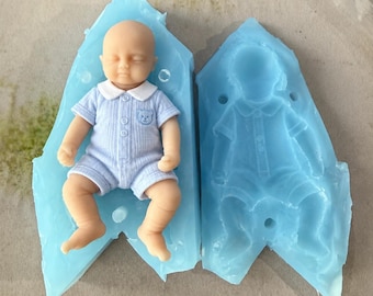 May include: A light blue silicone mold for a baby figurine, featuring a detailed impression of a sleeping infant wearing a collared romper with a small bear emblem on the chest, alongside a finished light blue baby figurine.