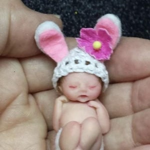 May include: A tiny, sleeping baby doll wearing a white crocheted hat with pink bunny ears and a pink flower. The doll is wearing a white diaper and has closed eyes.