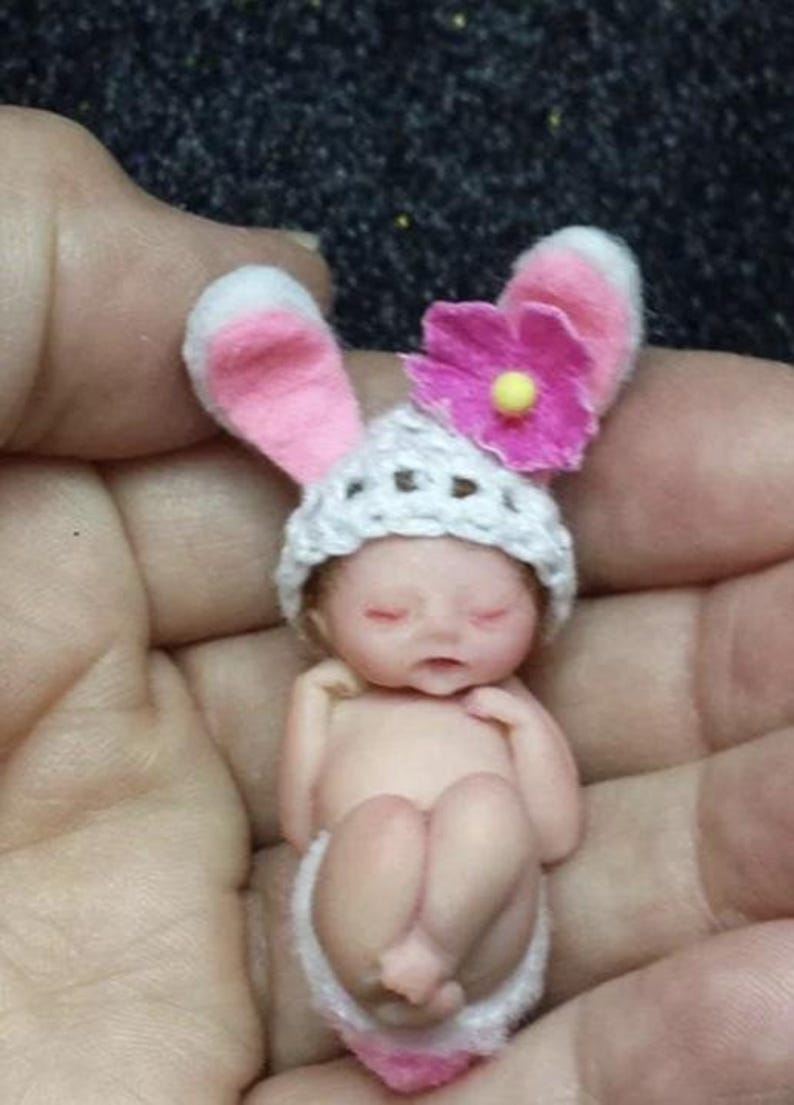 May include: A tiny, sleeping baby doll wearing a white crocheted hat with pink bunny ears and a pink flower. The doll is wearing a white diaper and has closed eyes.