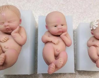 May include: Three silicone molds for making baby dolls. The molds are in different sizes and show a baby doll in each mold. The baby dolls are in sleeping poses.