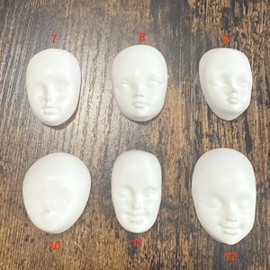 Puede incluir: Seis caras de muñecas blancas con diversas expresiones. Las caras son de forma ovalada con rasgos detallados, incluyendo ojos, narices y bocas. Las caras están numeradas 7, 8, 9, 10, 11 y 12.