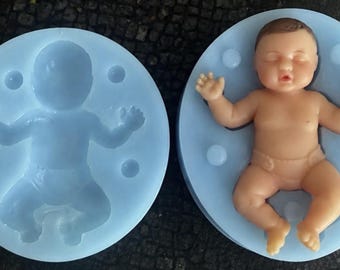May include: Two circular light blue silicone molds for crafting. One mold is empty, showing the negative impression of a sleeping infant. The other mold contains a realistic, flesh-toned, three-dimensional figure of a sleeping baby.