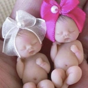 May include: Two small, sleeping baby dolls with bows on their heads. The doll on the left has a white bow and the doll on the right has a pink bow.