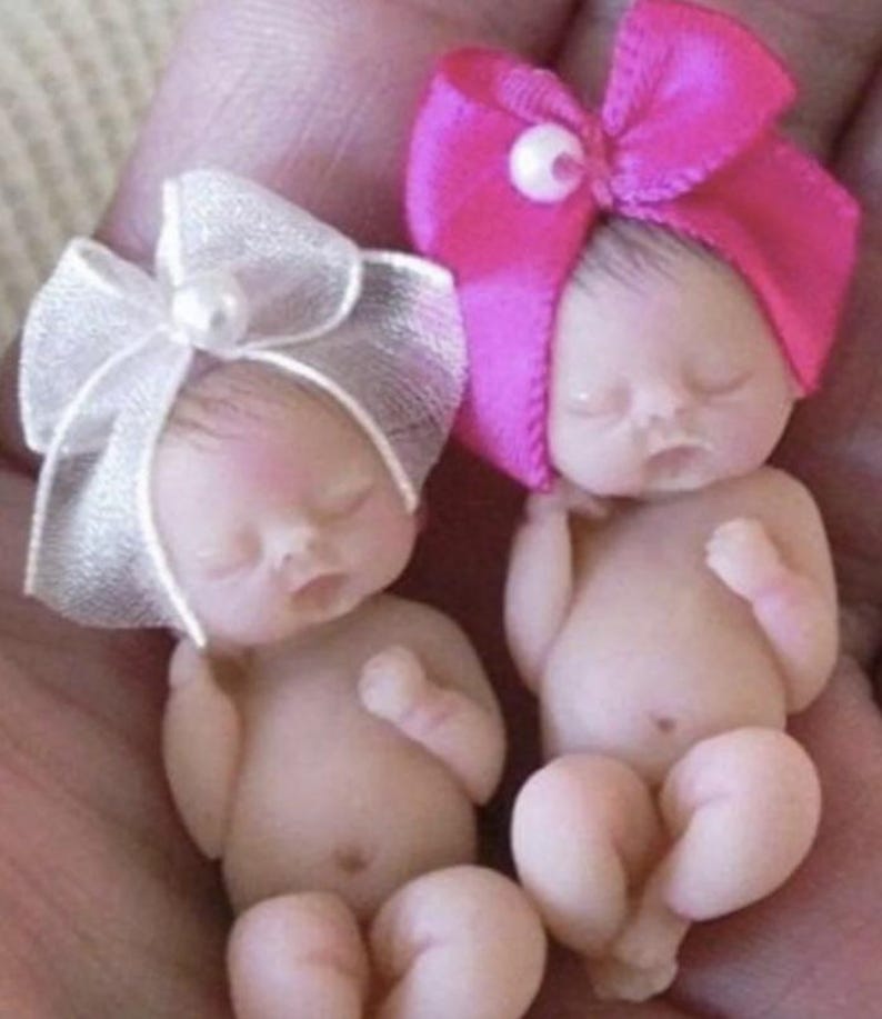 May include: Two small, sleeping baby dolls with bows on their heads. The doll on the left has a white bow and the doll on the right has a pink bow.