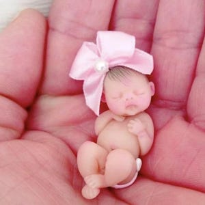May include: A tiny, sleeping baby doll with a pink bow and a pearl on top of the bow. The doll is wearing a diaper and is lying on its back with its arms crossed.