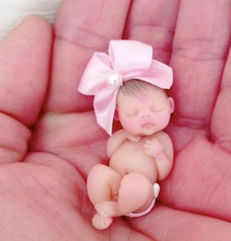 May include: A tiny, sleeping baby doll with a pink bow and a pearl on top of the bow. The doll is wearing a diaper and is lying on its back with its arms crossed.