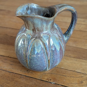 Op de afbeelding: Handgemaakte keramische kan met een bolvormig lichaam en een gebogen handvat. De kan heeft een glanzend glazuur in tinten blauw, bruin en paars. Het ontwerp omvat verticale groeven, die textuur toevoegen. Het is een decoratief item.