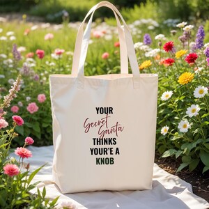 May include: A cream tote bag with the text "YOUR Secret Santa THINKS YOU'RE A KNOB" in burgundy and black. The bag has two handles. The background features a variety of colorful flowers.