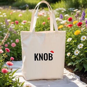 May include: A cream-coloured tote bag featuring the word "KNOB" in bold black letters, with a small red Santa hat icon. The bag has long handles and is set against a backdrop of colourful flowers.