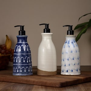May include: Three ceramic soap dispensers with black plastic pump tops. The left dispenser is dark blue with white arched patterns. The center dispenser is solid off-white with a brown base. The right dispenser is white with blue brushstroke patterns.