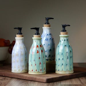 May include: Four ceramic soap dispensers with black pumps and cork stoppers. The dispensers are in shades of white, blue, and green with decorative patterns. They are arranged on a wooden tray.
