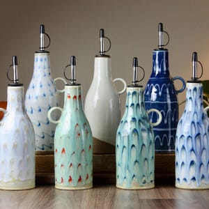 May include: A collection of ceramic oil bottles with stainless steel spouts and small handles. The bottles are in various colors, including white, blue, and green, with decorative patterns. They are arranged on a wooden surface.