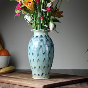 May include: A tall, blue and white ceramic vase with a ruffled rim, filled with colorful flowers. The vase has a unique pattern of blue teardrop shapes. The arrangement sits on a wooden cutting board, with fruit and a basket in the background.