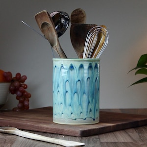 May include: A ceramic utensil holder with a blue and white drip glaze design. The holder contains wooden spoons, a whisk, and other kitchen tools. It sits on a wooden cutting board, with a wooden spoon in the foreground.