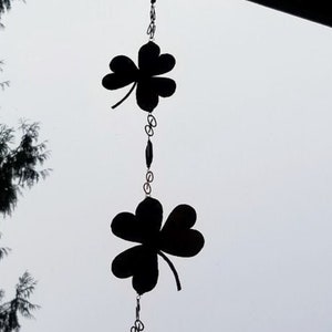 May include: A black metal wind chime with three shamrock shapes and a bell at the bottom. The wind chime is hanging from a chain.