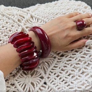 May include: A hand wearing a burgundy ring and two bracelets. One bracelet is a solid, rounded bangle, and the other is a multi-layered, textured design. The jewelry is a deep red color with a glossy finish.