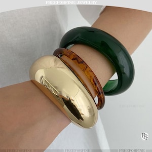 Può includere: Tre braccialetti in diversi stili e colori sono impilati su un polso. Un bracciale largo e dorato è accanto a un braccialetto traslucido color ambra. Un braccialetto arrotondato verde scuro completa il set.