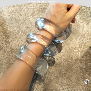 Op de afbeelding: Drie heldere, gladde, afgeronde armbanden gedragen op de onderarm. De armbanden zijn transparant en hebben een dikke, solide constructie met een gepolijste afwerking. De tekst FREEFORFINE | JEWELRY is bovenaan en FFF onderaan zichtbaar.