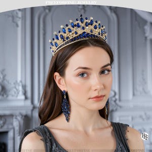 Op de afbeelding: Een goudkleurige kroon en bijpassende oorbellen, beide versierd met diepblauwe edelstenen. De kroon heeft een gedetailleerd ontwerp met afwisselend grote en kleine stenen. De oorbellen vallen in een cascade naar beneden, waardoor een samenhangende sieradenset ontstaat. De afbeelding is van FREEFORFINE JEWELRY.