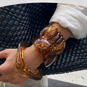 May include: A set of three amber-colored resin bracelets with a glossy, translucent finish. The bracelets feature a smooth, rounded design with varying thicknesses, worn together on the wrist. The text FREEFORFINE JEWELRY appears at the top.