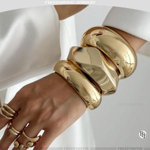 May include: Gold-toned jewelry set featuring multiple chunky bracelets and rings. The bracelets have a smooth, polished finish, while the rings showcase unique designs. The jewelry is displayed on a hand, with the brand name "FREEFORFINE JEWELRY" visible.