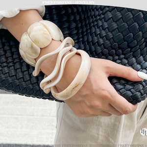 May include: A collection of cream and ivory-toned bracelets and bangles. The chunky bracelet and bangles are worn on a wrist next to a black woven handbag. The image has the text "FREEFORFINE JEWELRY" at the top and bottom.