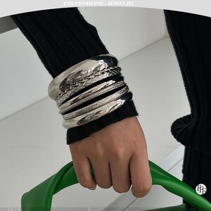 May include: A stack of six silver-toned metal bracelets worn on the wrist. The set includes a smooth wide bangle, a braided chain, a thin polished band, a textured chain, a flat etched band, and a second smooth wide bangle in a silver finish.