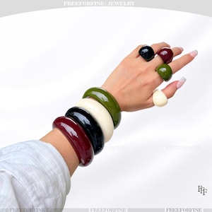 Pode incluir: Uma coleção de joias de resina coloridas e grossas. A imagem apresenta uma variedade de anéis e pulseiras em tons de preto, bordô, branco, verde-oliva e verde escuro. As joias têm um acabamento brilhante e um design moderno e arrojado.