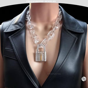 May include: A clear acrylic chain necklace with a padlock pendant. The chain links are large and oval-shaped. The padlock is transparent with a gold-coloured interior. The necklace is worn over a black leather vest.