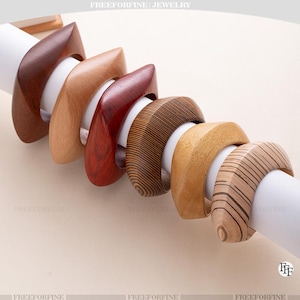 May include: Six wooden bracelets in shades of brown, from light tan to dark mahogany. Each bracelet has a square, curved design. The bracelets are displayed on a white cylindrical form. The text "FREEFORFINE JEWELRY" is at the top.