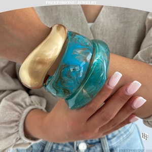 May include: A set of three chunky bangles worn together. One bangle is a solid, smooth matte gold tone. The other two bangles feature a translucent, swirling marbled pattern in shades of bright turquoise, deep teal, and hints of white throughout.