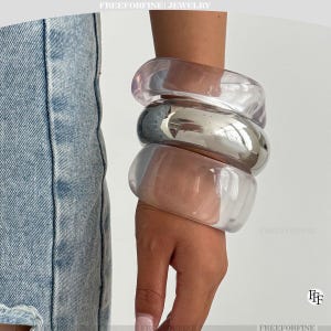Può includere: Tre braccialetti spessi in acrilico trasparente e un braccialetto argentato sono impilati su un polso. I braccialetti trasparenti hanno una sfumatura leggermente rosata. I gioielli sono ambientati su uno sfondo bianco. Il testo "FREEFORFINE JEWELRY" è in alto.