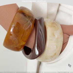 Op de afbeelding: Vier dikke harsarmbanden in bruin- en crèmetinten. De armbanden hebben een gemarmerd effect en een glanzende afwerking. De sieraden worden getoond op de arm van een persoon. De merknaam "FREEFORFINE JEWELRY" staat bovenaan.