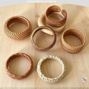 May include: Six circular bracelets crafted from woven rattan material. Five bracelets feature a natural brown finish, while one bracelet displays a lighter, cream-colored tone. The items are arranged in a cluster, with one bracelet resting on a small wooden base.