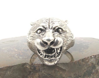 May include: A silver ring featuring a detailed, snarling tiger head design. The ring is displayed on a rough, brown rock.
