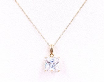 May include: A gold necklace featuring a square, clear gemstone pendant. The pendant is set in a gold bezel and suspended from a fine gold chain. The necklace is shown against a white backdrop.