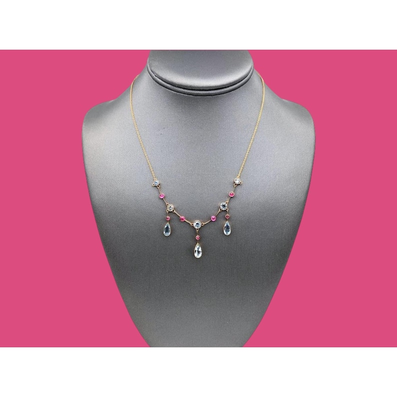 May include: A gold necklace with a delicate design featuring alternating pink and clear gemstones. The necklace has a teardrop pendant with a clear gemstone.