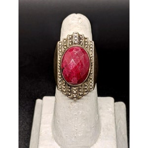 May include: A silver ring with a large, oval-shaped, faceted ruby gemstone. The ring has a decorative band with small, white beads.