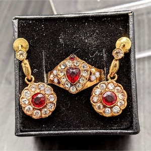 Może przedstawiać: Zestaw złotych ozdób z czerwonymi, sercowatymi kamieniami szlachetnymi otoczonymi przez przezroczyste cyrkonie. Zestaw zawiera pierścionek, parę kolczyków i dwa wisiorki.