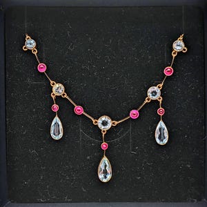 May include: A gold necklace with pink and clear stones. The necklace has a teardrop design with three dangling stones.