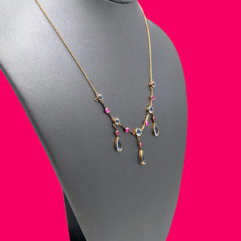 May include: A gold necklace with a delicate chain and alternating blue and pink gemstones. The necklace features three teardrop-shaped pendants.