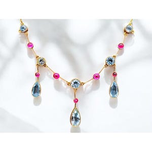 May include: A gold necklace with a delicate chain and alternating pink and blue gemstones. The necklace features three teardrop-shaped blue gemstones hanging from the chain.