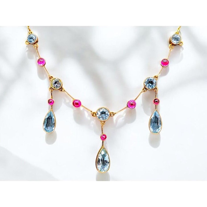 May include: A gold necklace with a delicate chain and alternating pink and blue gemstones. The necklace features three teardrop-shaped blue gemstones hanging from the chain.