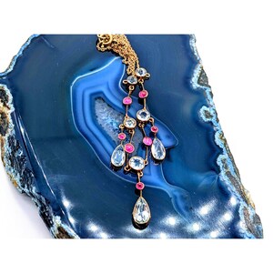 May include: A gold necklace with a teardrop-shaped pendant and multiple smaller teardrop-shaped stones. The necklace is adorned with pink and clear stones.