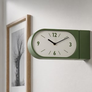 Könnte beinhalten: Eine moderne, olivgrüne Wanduhr mit rechteckigem Design. Das Zifferblatt ist weiß mit schwarzen Zeigern und Ziffern. Die Uhr ist an einer weißen Wand montiert, mit einem gerahmten Schwarz-Weiß-Bild im Hintergrund.