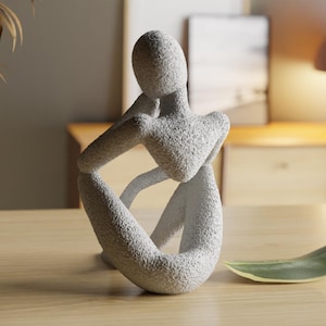 Pode incluir: Uma escultura abstrata texturizada, cinza claro, de uma pessoa em pose sentada, com os braços apoiados nos joelhos. Uma folha verde e branca repousa sobre a superfície de madeira. A escultura é um acessório decorativo para casa.