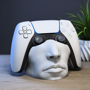 Könnte beinhalten: Ein weiß-schwarzer PlayStation 5-Controller liegt auf einer weißen Büste eines Gesichts. Der Controller hat blaue Akzente und das Standard-Tastenlayout. Eine kleine grüne Pflanze in einem weißen Topf steht im Hintergrund.